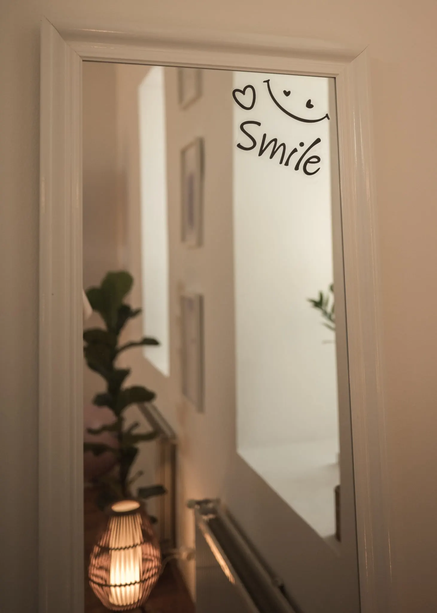 Ajtóra szerelt tükör „Smile” matricával, alatta rattan lámpa és növény